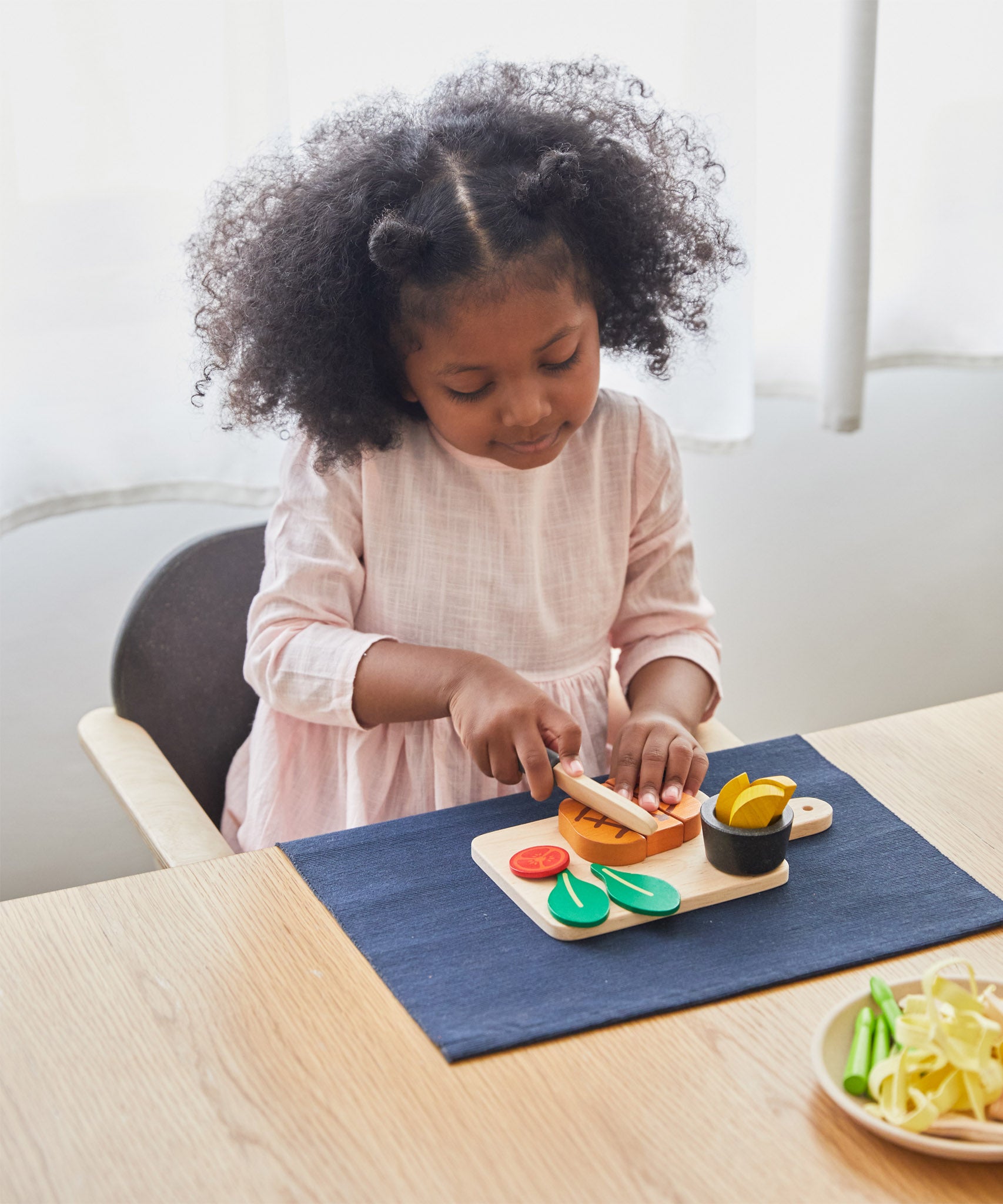 A girl sat by the table and cutting through the steak on the cutting board from the Plan Toys wooden steak set and available at Babipur.