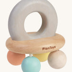 PlanToys Pastel Bell Rattle, is a beautiful wooden baby toy. The rattle comes in the shape of a bell, with a large soft grey wooden ring on top for easy gripping. At the bottom of the ring is a horizontal wooden oval, and attached to the wooden oval are four pastel coloured balls in blue, white, peach and yellow. The image is on a cream background