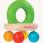 PlanToys Bell Rattle, is a beautiful wooden baby toy. The rattle comes in the shape of a bell, with a large green wooden ring on top for easy gripping. At the bottom of the ring is a horizontal wooden oval, and attached to the wooden oval are four bright coloured balls in blue, orange, red and yellow. The image is on a cream background