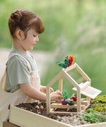 A PlanToys wooden blooming greenhouse standing on soil with a child sorting different veg pieces
