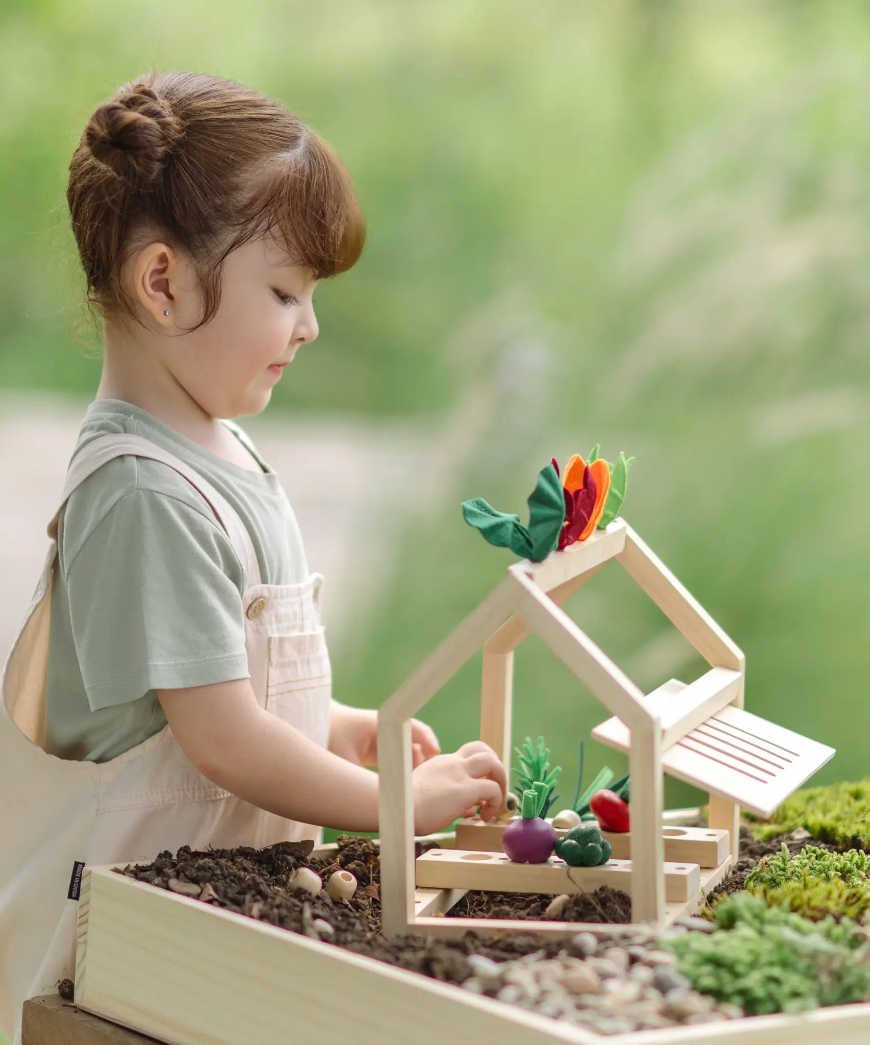 A PlanToys wooden blooming greenhouse standing on soil with a child sorting different veg pieces