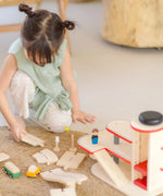 Child holding on loose PlanToys wooden road pieces next to a car park from the city transit rail road playset