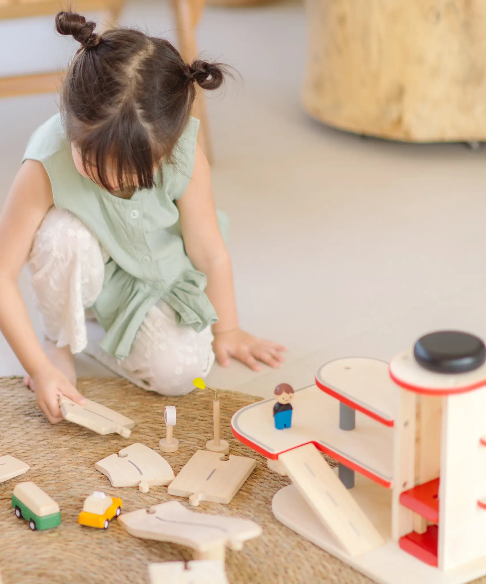 Child holding on loose PlanToys wooden road pieces next to a car park from the city transit rail road playset