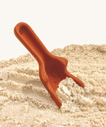 The orange scoop from the PlanToys Sand Play Set. The scoops has texture dots on the front and the back, and the scoop is digging in the sand