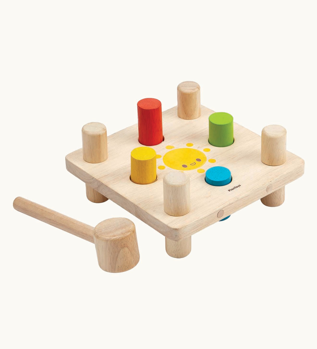 The Plan Toys Wooden Hammer Peg  toy, is a classic wooden toy hammer bench with colourful pegs for babies and toddlers. The bench is square in shape with a smiling sun in the middle, and has a red, yellow, blue and green peg. There is also a wooden hammer next to the peg board. The image is on a cream background.