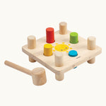 The Plan Toys Wooden Hammer Peg  toy, is a classic wooden toy hammer bench with colourful pegs for babies and toddlers. The bench is square in shape with a smiling sun in the middle, and has a red, yellow, blue and green peg. There is also a wooden hammer next to the peg board. The image is on a cream background.