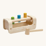 The hammer/mallet is in front of the PlanToys Pounding Bench. The hammer/mallet has one flat side and one rounded side. The image is on a cream background