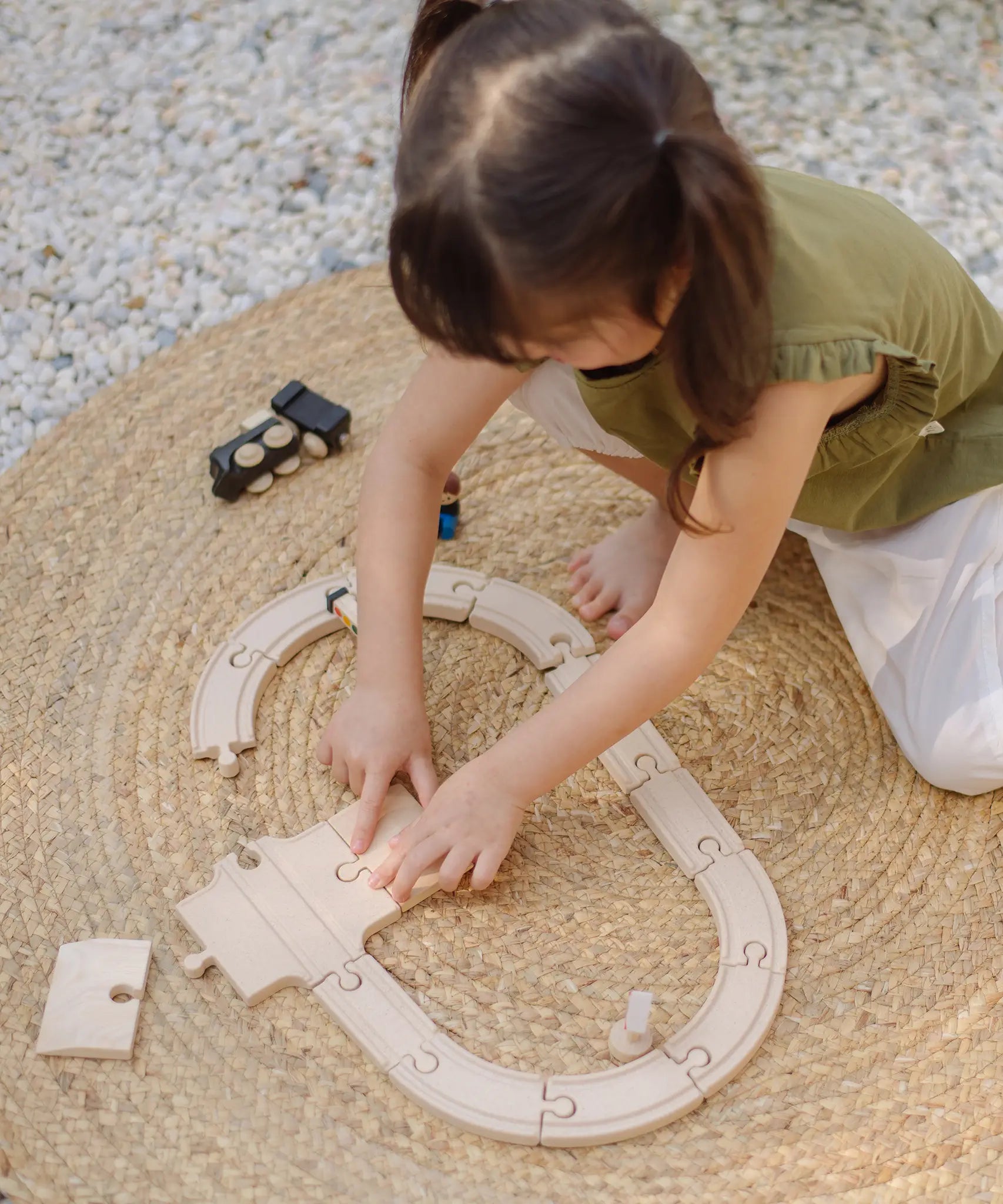Child assembling wooden rail pieces from the PlanToys my first train set on a woven rug in the garden