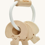 PlanToys Wooden Baby Key Rattle is a fun sensory toy for little ones. This toy is great for teething and soothing sore gums. There is a circular white rope with a wooden bead at the top. Attached to the rope are three, natural wooden keys, all with smiling faces. The image is on a cream background.