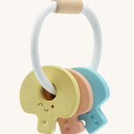PlanToys Wooden Baby Key Rattle is a fun sensory toy for little ones. This toy is great for teething and soothing sore gums. There is a circular white rope with a wooden bead at the top. Attached to the rope are three, colourful pastel wooden keys, all with smiling faces and dots to correspond to the key. The yellow key has one dot, the peach key has two dots, and the blue key has three dots. The image is on a cream background.