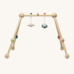 The PlanToys Play Gym in Orchard colours (natural wood, green, blue, brown and beige) with the rocket and planet sensory toys attached to the top using a blue and a red peg hook. This image is front on, on a cream background