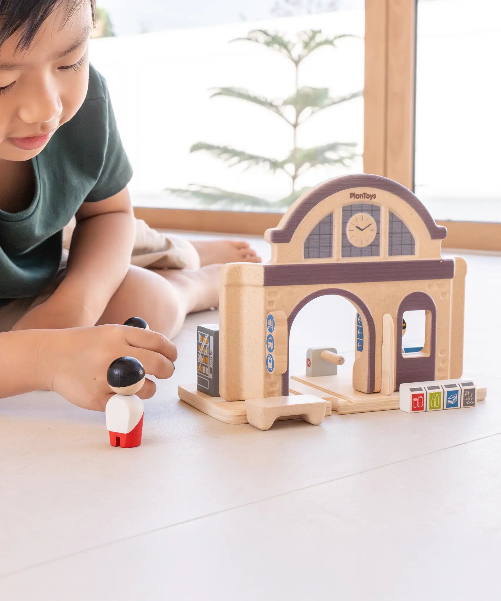 Child holding wooden people figures next to a PlanToys train station 