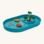 PlanToys Water Play Set is made from natural rubber and is great for sensory water play. The image shows a blue tray filled with water, with 3 fish floating on the water, a figure in a PlanToys boat, a boat tied to a brown pole and a figure stood on a wood log with palm trees to give a raised island effect. The image is on a cream background