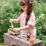 Child holding a wooden spade with a PlanToys wheelbarrow filled with soil 