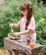 Child holding a wooden spade with a PlanToys wheelbarrow filled with soil 