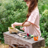 Child holding a wooden watering can from the garden wheelbarrow set with other garden tools on a wooden bench