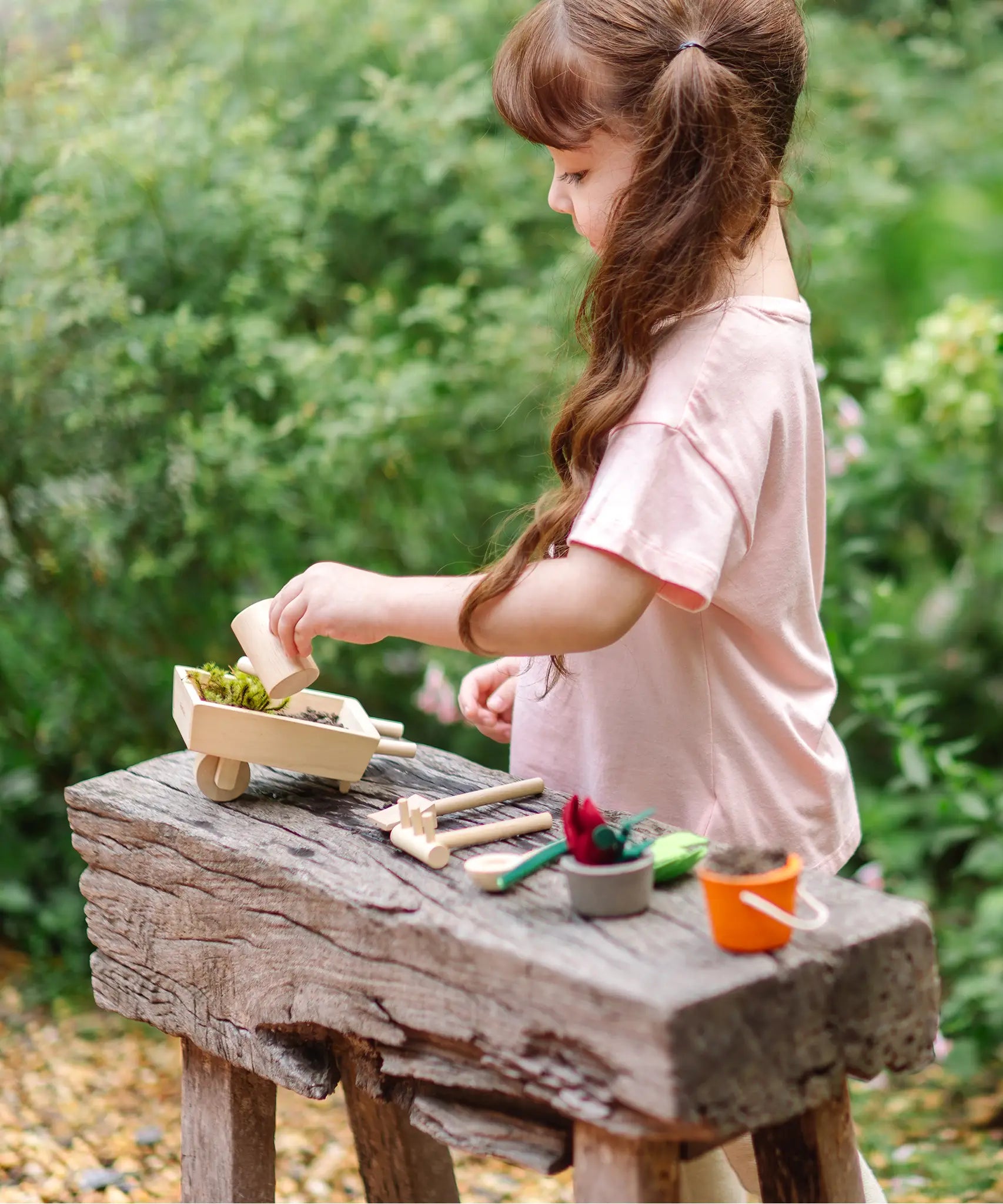 Child holding a wooden watering can from the garden wheelbarrow set with other garden tools on a wooden bench