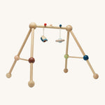 The PlanToys Play Gym Orchard is a fun, sensory baby gym made from wood. This set includes a rocket and a planet toy attached to the top of the baby gym using a red and a blue peg. This is a side view image on a cream background.