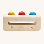 A side view of the PlanToys Hammer Balls set showing the yellow, rd, and blue ball resting on top of the hammer board. The image is on a cream background