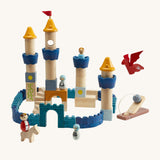 The PlanToys Castle Building Blocks in Orchard colours, made into a colourful castle display. The set comes with castle wall blocks in shades of blue, blocks to make castle towers and turrets, little people figures, a horse, a dragon and a catapult to defend the castle. The image is on a cream background