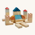 PlanToys Creative Blocks in the colourwave Orchard, are a set of building blocks in various shapes and colours which include natural coloured blocks, coral pink, blue, red, grey and mustard. The image is on a cream background