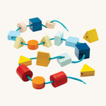PlanToys children's wooden geo lacing blocks laid out on a cream background. The image shows wooden bead blocks in various colours and shapes, threaded on a light blue shoe string