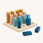 The PlanToys Geometric Peg Board is a fun, educational, shapes game for children. The image shows a PlanWood square base, with 16 shape cutouts, 4 circles, 4 rectanges, 4 triangles and 4 square. There are wooden blocks in each cut out with larger blocks at the back, going down to smaller blocks in the front. There is a rectangle and square block in front of the play set, and the colour of the blocks are navy blue, light blue, peach and orange/yellow. The image is on a cream background.