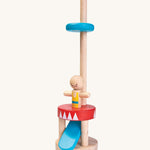The PlanToys Jumping Acrobat is a magical toy which consists of a wooden peg acrobat with a yellow and blue top, a smiling face with a mustache, a red and white base, a blue push pedal underneath the base, and a blue disk on a wooden pole to catch the figure when it pops up. The image is on a cream background
