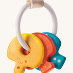 PlanToys Baby Key Rattle is a fun sensory toy for little ones. This toy is great for teething and soothing sore gums. There is a circular white rope with a wooden bead at the top. Attached to the rope are three, colourful wooden keys, all with smiling faces and dots to correspond to the key. The yellow key has one dot, the red key has two dots, and the blue key has three dots. The image is on a cream background.