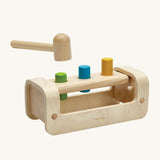 PlanToys Wooden Pounding Bench, has a curved, semi-circular base with a rectangle peg board with 3 holes, to allow for 3 wooden pegs to go through. The base allows for movement of the toy in a rocking motion. In this image there is a wooden hammer/mallet above the pegs in the peg board and the peg colours are green, blue and yellow. This image is on a cream background