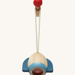 A closer look at the rocket toy from the PlanToys Play Gym in Orchard colours, on a red ball hook. The rocket is made from natural wood showing the wood grain and a light blue band at the bottom, dark blue rocket fins, and there is also a red ball inside. The image is on a cream background