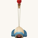 A closer look at the rocket toy from the PlanToys Play Gym in Orchard colours, on a red ball hook. The rocket is made from natural wood showing the wood grain and a light blue band at the bottom, dark blue rocket fins, and there is also a red ball inside. The image is on a cream background