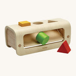 The PlanToys Shape and Sort is made from natural wood, and has a semi circle wooden base to allow for movement when passing the shapes through the holes. There is a slanted section inside the toy to allow the wooden shapes to fall out. The shapes are a yellow square, a green circle and a red triangle. The image is on a cream background