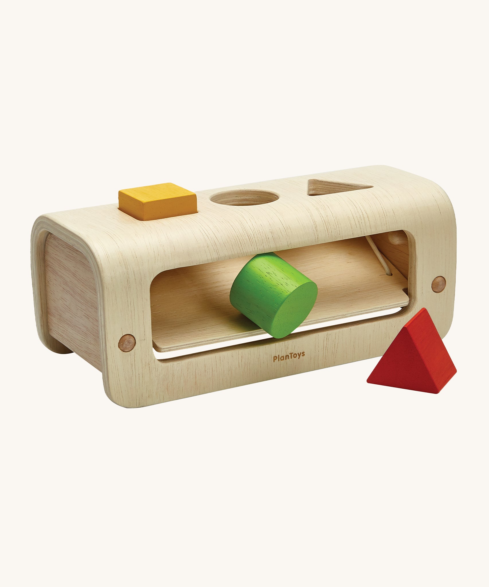 The PlanToys Shape and Sort is made from natural wood, and has a semi circle wooden base to allow for movement when passing the shapes through the holes. There is a slanted section inside the toy to allow the wooden shapes to fall out. The shapes are a yellow square, a green circle and a red triangle. The image is on a cream background