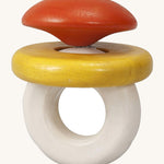 This PlanToys Baby Rattle, is in the shape of a ring, with an orange/red diamond jewel shape on top, a white ring attached for easy grasping, and a horizontal yellow ring which moves. The image is on a cream background