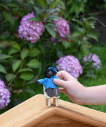 A child holding a doll from the PlanToys doll house family set in their hand. The child is placing it on a wooden toy seat.