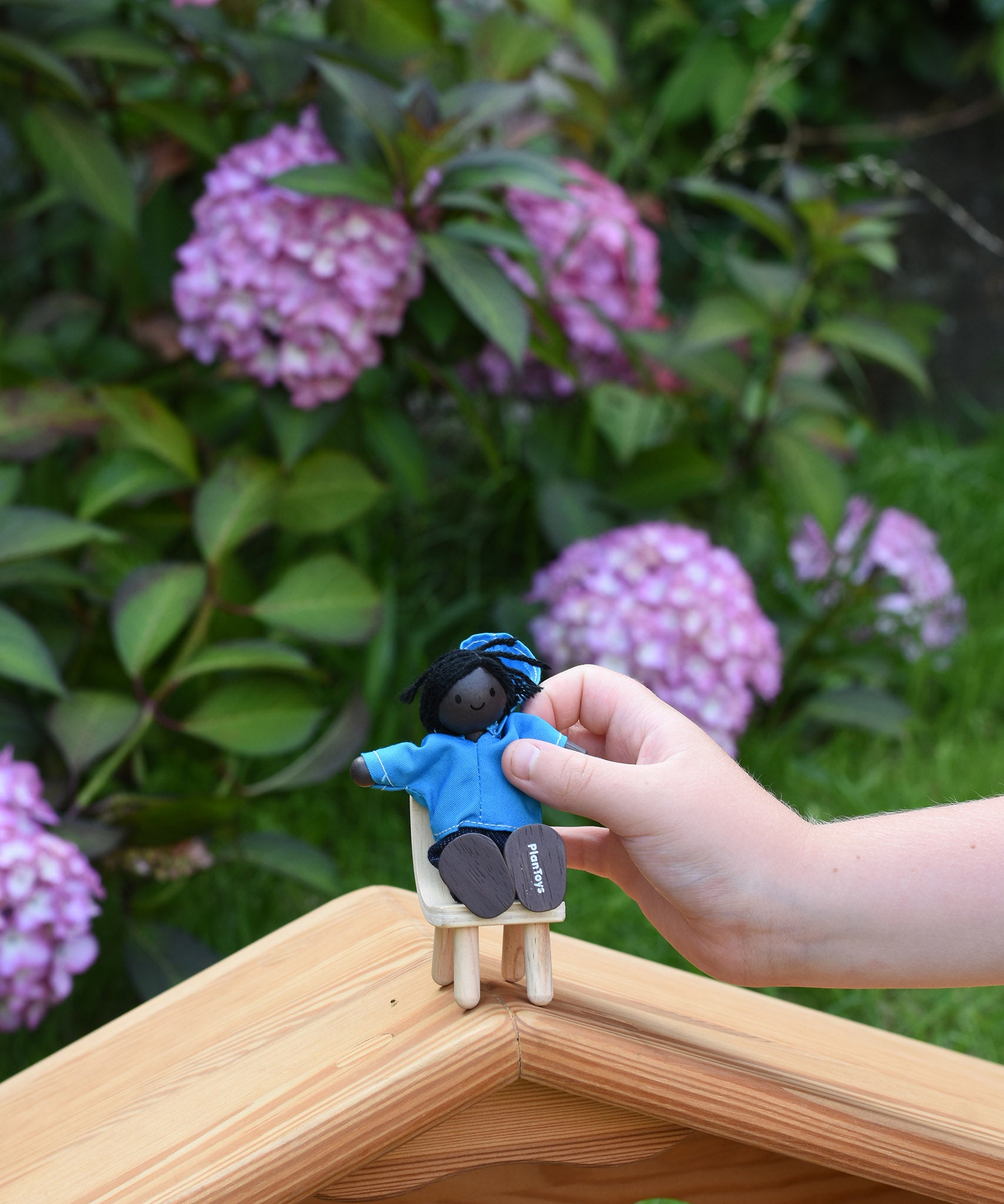 A child holding a doll from the PlanToys doll house family set in their hand. The child is placing it on a wooden toy seat.