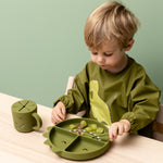 Child wearing the Trixie Animal Friends Waterproof Long Sleeve Bib, green, Mr Dino, with matching tableware.