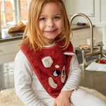 A child sat in a kitchen, wearing the Roommate Organic Cotton Baby Bib - Red Panda