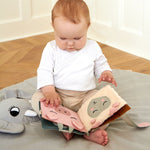 A baby sat on a Roommate playmat, playing with the Roommate Baby Nursery Book - Country Life on its lap. The fabric pages show a pig and a sheep