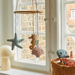 The Roommate Sealife Hanging Mobile hung in front of a window with Roommate toys in a basket and a mushroom lamp on the window ledge