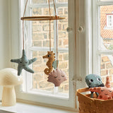The Roommate Sealife Hanging Mobile hung in front of a window with Roommate toys in a basket and a mushroom lamp on the window ledge