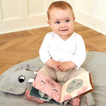 A baby sat on a Roommate playmat, with the Roommate Baby Nursery Book - Country Life on its lap. The fabric pages show a pig and a sheep