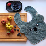 A top down view of the Roommate Organic Cotton Baby Bib - Koala on a white table top, half placed on a wooden chopping board with fruit on it. Above the chopping board is a black bowl with fruit inside