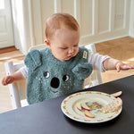 A child sat at a table whilst eating, wearing the Roommate Organic Cotton Baby Bib - Koala