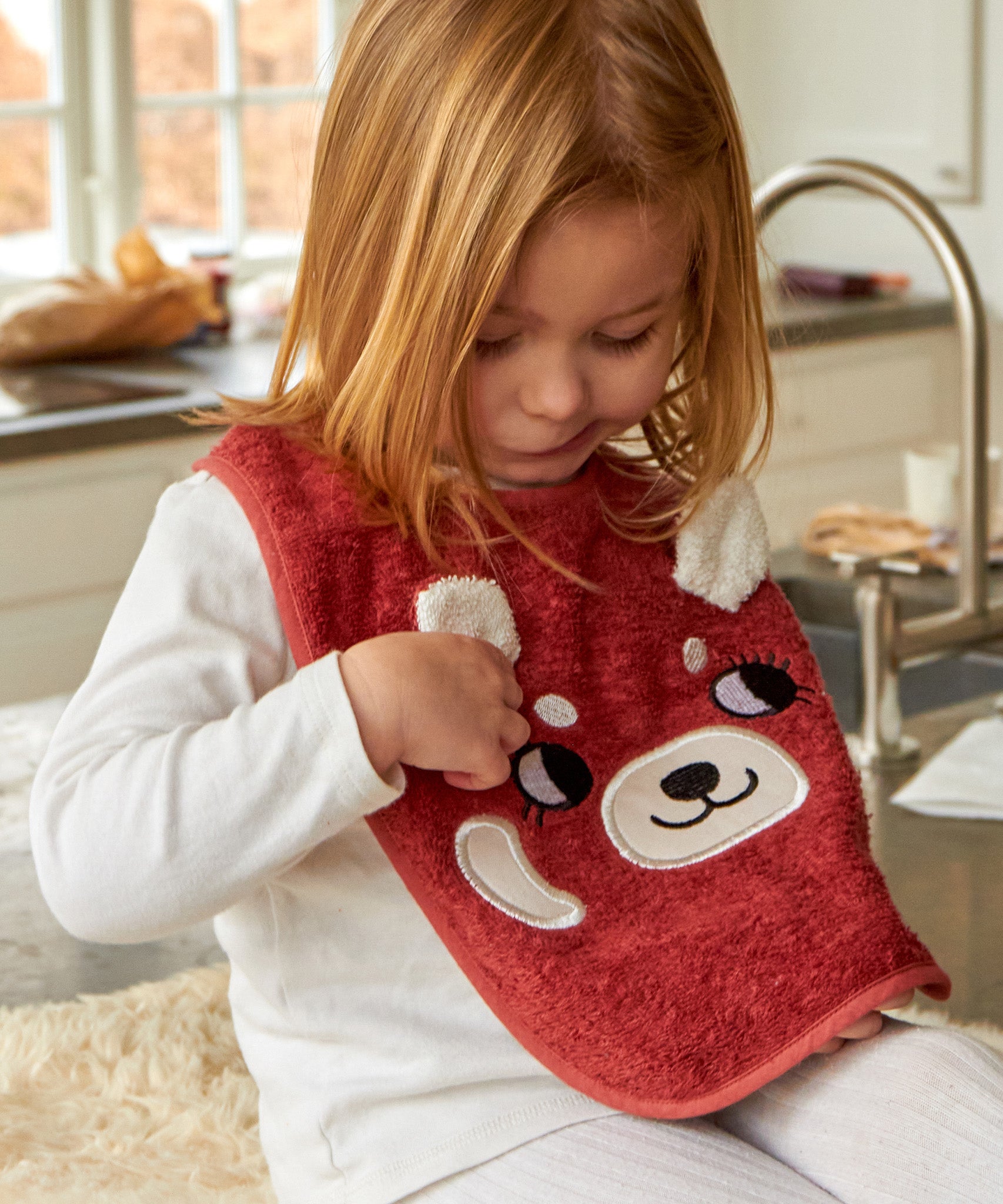 A child sat in a kitchen looking down at the Roommate Organic Cotton Baby Bib - Red Panda
