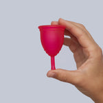 A person holding the Red Silicone, BPA free and latex free Ruby Cup in between their fingers, on a light grey background