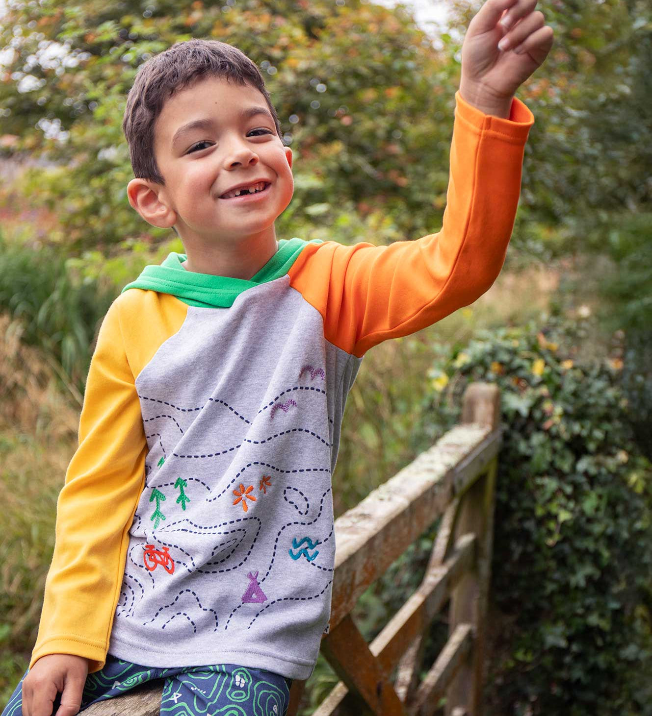 A child smiling, sitting on a gate wearinga Frugi Compass Organic Cotton Hooded Top. 