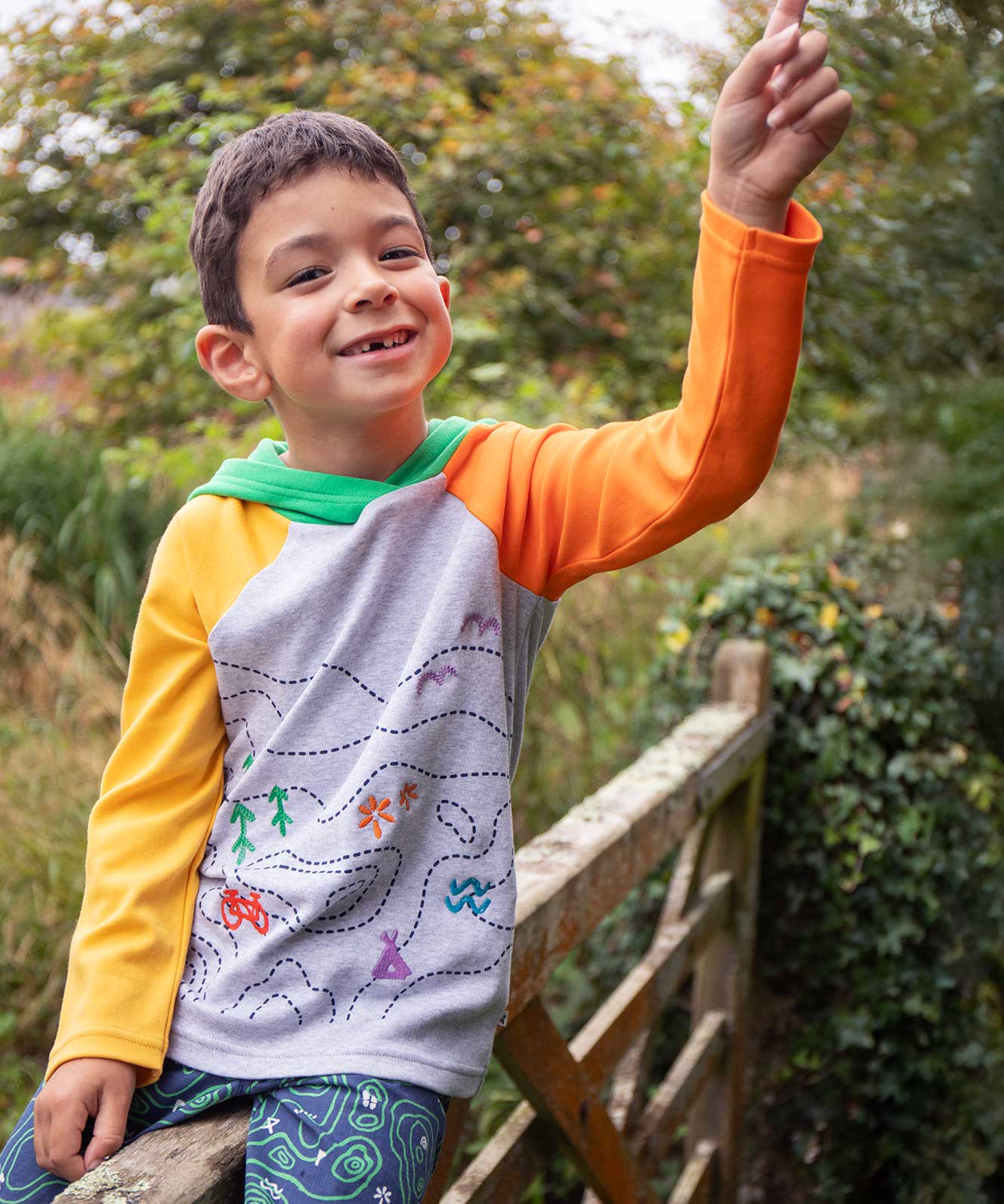 A child smiling, sitting on a gate wearinga Frugi Compass Organic Cotton Hooded Top. 