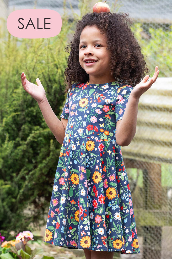Child in an allotment wearing Frugi Sunshine Skater Dress, navy in a floral Potager Garden print, made from GOTS organic cotton, to represent Frugi Sale at Babipur.
